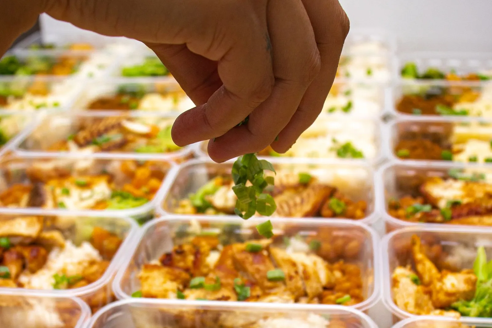 A cook sprinkling herbs over rows of packed meal prep containers with chicken and rice